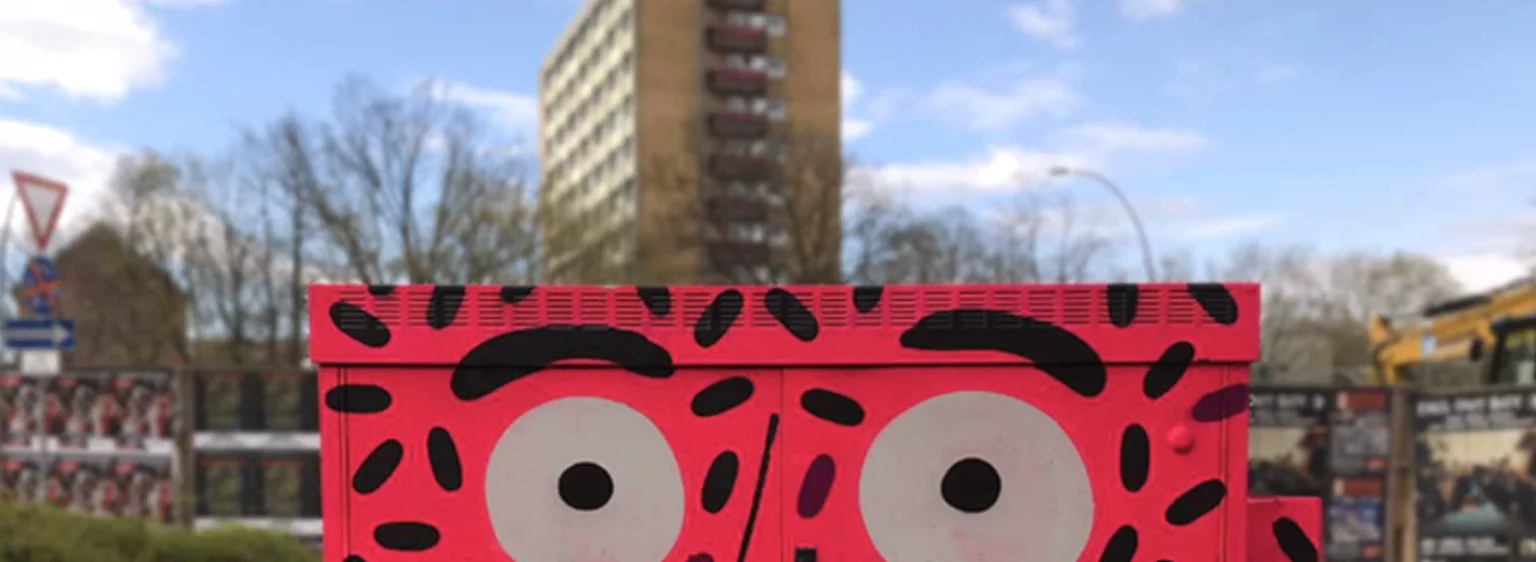 Fertig bemalter Stromkasten mit pinkem Gesicht, Himmel und Hochhaus im Hintergrund.