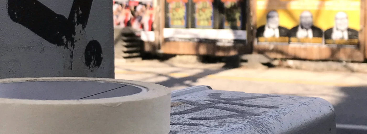 Klebebandrolle liegt auf dem Stromkasten, im Hintergrund unscharf Plakatwand und Baustelle.