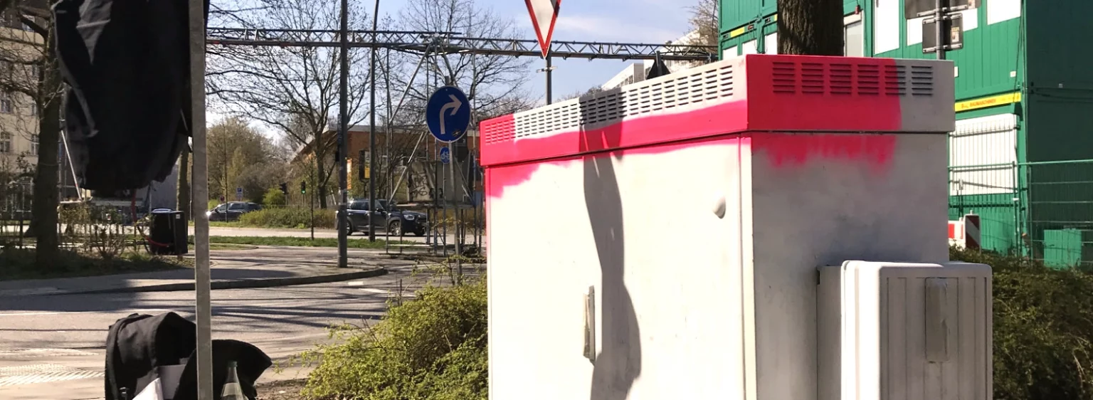 Stromkasten halb bemalt, seitliche Ansicht, Baustellencontainer im Hintergrund.