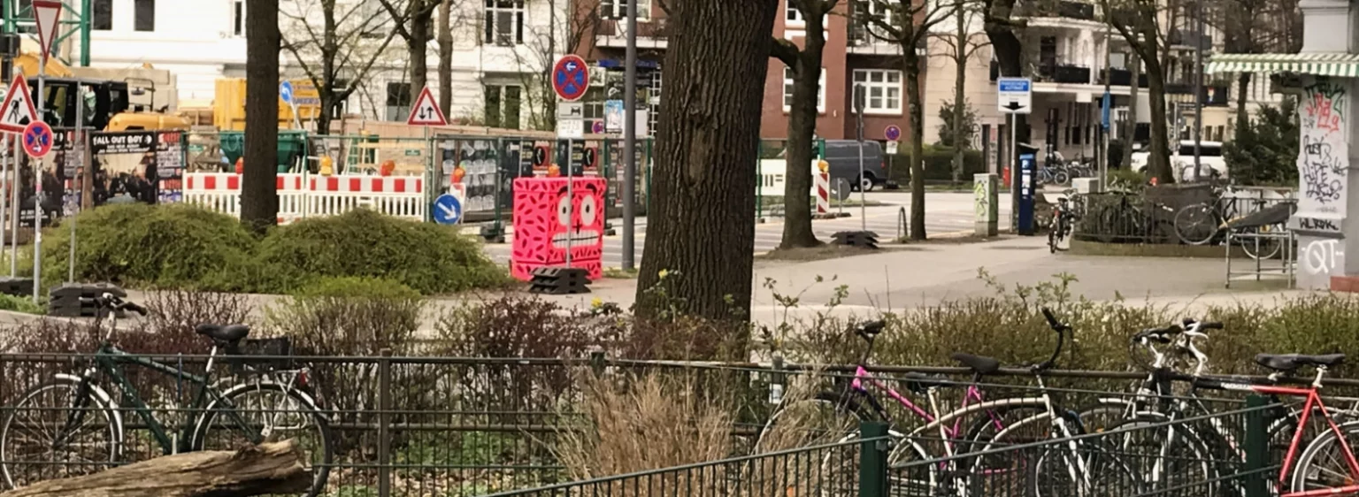 Stromkasten aus der Entfernung, im Vordergrund Fahrräder, Baustellenschilder und Bäume.