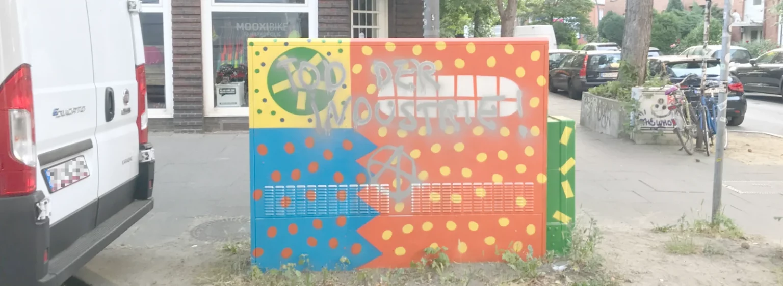 Ein bemalter Stromkasten mit farbenfrohem, grafischem Motiv, das inzwischen durch spontane Ergänzungen aus der Nachbarschaft erweitert wurde; ein urbanes Kunstwerk im Alltag, gestaltet von Fiete Rakete.