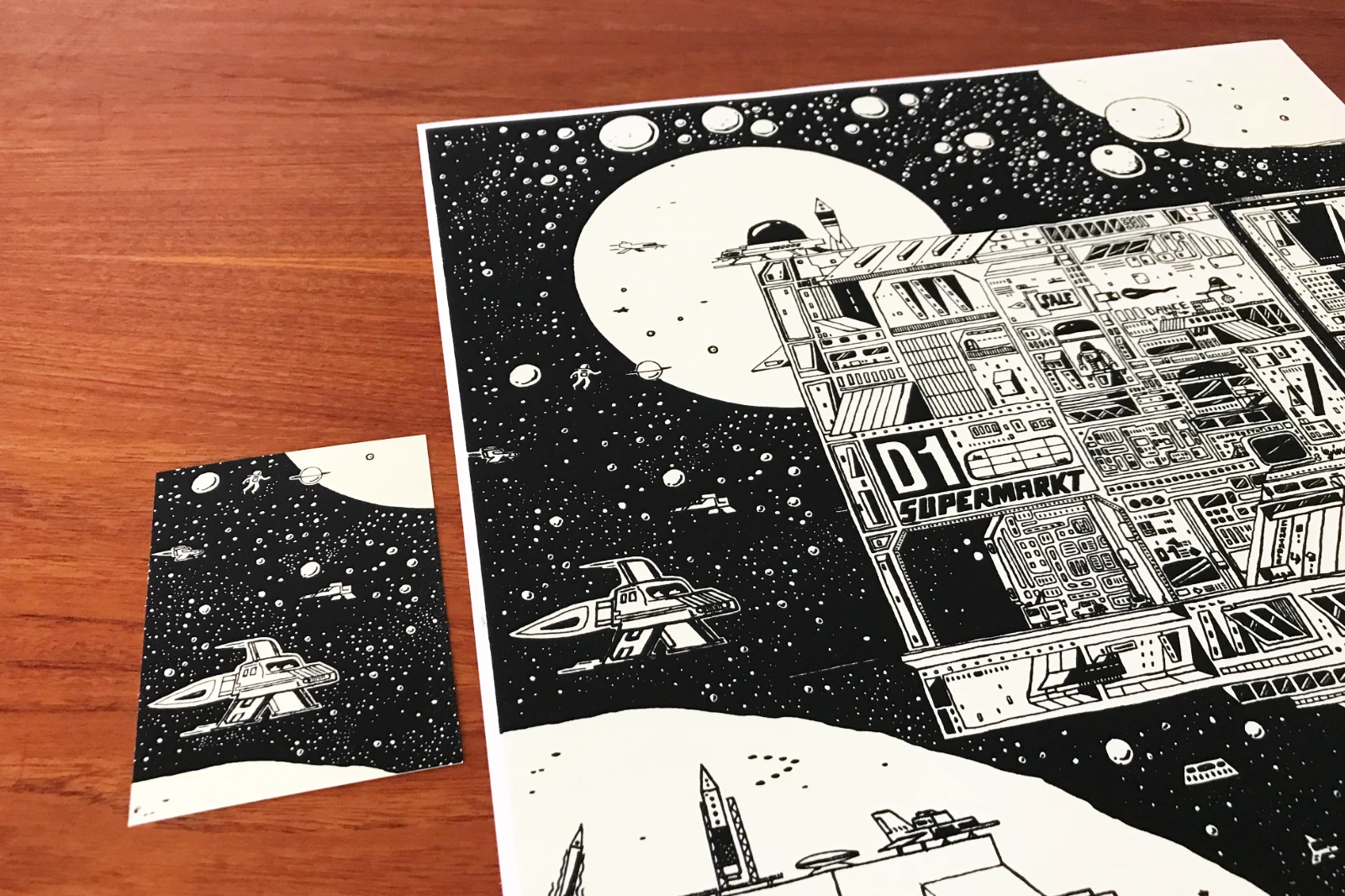 Poster und Postkarte des limitierten Siebdruckmotivs „Krom City“ von Fiete Rakete, gemeinsam auf einem Tisch liegend; das Poster ist nur teilweise zu sehen, die Postkarte vollständig.